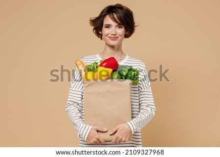 Young smiling caucasian cheerful happy fun vegetarian woman 20s in casual clothes hold paper bag with vegetables after shopping look camera isolated on plain pastel beige background studio portrait Royalty-Free Stock Photo #2109327968