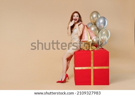 Full size surprised young woman in dress near balloons sit on big red gift present box hold shopping package bags purchases isolated on plain pastel beige background Celebrating birthday party concept