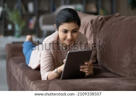 Cheerful young Indian woman using digital computer tablet, lying on comfortable sofa, choosing goods shopping in internet store, communicating distantly in social network, playing games online.