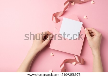 First person top view photo of valentine's day decor young woman's hands holding open pink envelope with paper card hearts and pink silk curly ribbon on isolated pastel pink background with copyspace