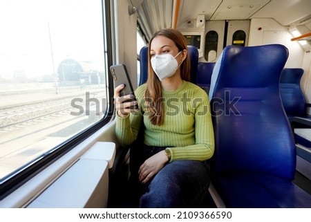 Young woman wearing medical mask relaxing in train seat while using smartphone app. Business woman enjoying view texting on mobile phone. Travel safety lifestyle. Royalty-Free Stock Photo #2109366590