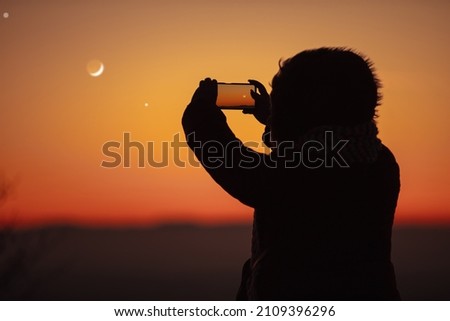 Woman taking photo with smartphone of evening sky with crescent Moon, stars and planets.