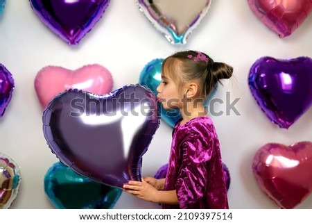 A girl in a purple dress kisses a heart-shaped balloon on the background.  Decorations for the Valentine's Day party. Colorful foil balloons decorate the wall