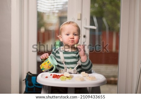 Caucasian blonder toddler male sitting in high chair holding drink and eating lunch.