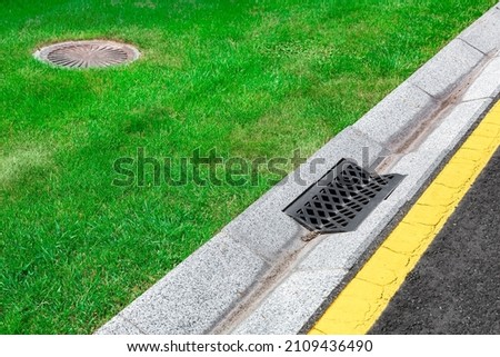 gray gutter of a stormwater drainage system on the side of asphalt road with yellow markings and sewer septic tank in green lawn, concrete drainage ditch with iron grate. Royalty-Free Stock Photo #2109436490