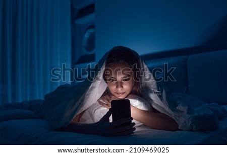 Young female lying under blanket on bed and watching horror movie on mobile phone in dark bedroom in late evening Royalty-Free Stock Photo #2109469025
