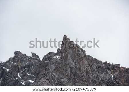 Gloomy mountain landscape with high sharp rockies with snow in gray rainy sky. Closeup of sharp rocks and peaked top in gray cloudy sky. Bleak overcast scenery with mountain range with pointy peak. Royalty-Free Stock Photo #2109475379
