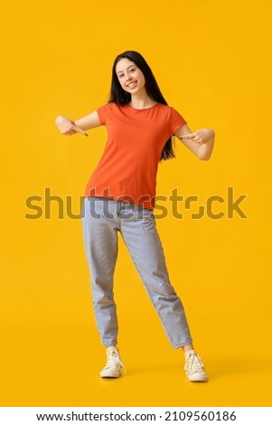 Beautiful teenage girl pointing at orange t-shirt on yellow background