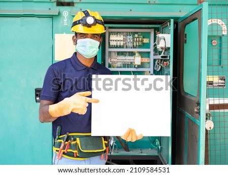 Electrician with meidcal face mask and safety electrician with meidcal face mask and safety precautions holding empty sign board by looking at camera - concept of advertisement,promotion and job offer Royalty-Free Stock Photo #2109584531