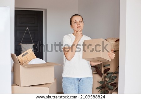 Indoor shot of pensive woman wearing white casual style T-shirt and jeans standing holding cardboard box with belongings, looking away, holding chin, thinking how to furnish the interior.