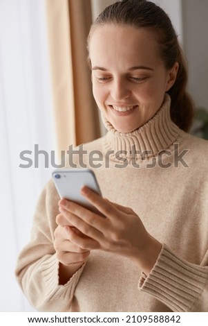 Portrait of delighted young adult woman with ponytail hairstyle wearing beige casual style sweater using mobile phone, writing post for social networks or watching photos.