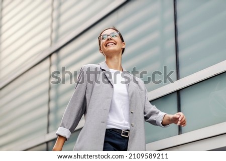 Low angle view of young, smiling businesswoman dressed elegantly passing by windows and going to her office. A successful businesswoman going to her workplace Royalty-Free Stock Photo #2109589211