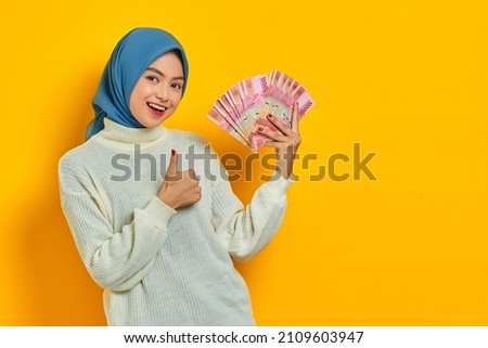 Smiling beautiful Asian Muslim woman in white sweater showing cash money in Indonesian rupiah banknotes, gesturing thumbs up isolated over yellow background. ​People religious lifestyle concept