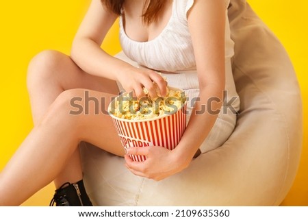 Young woman with tasty popcorn sitting in beanbag chair on yellow background