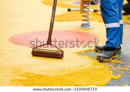 Worker leveling and pressing rubber coating for playgrounds with roller, mason hand spreading soft rubber crumbs. Outdoor soft coating and floor covering for sports. Selective focus. Royalty-Free Stock Photo #2109648719