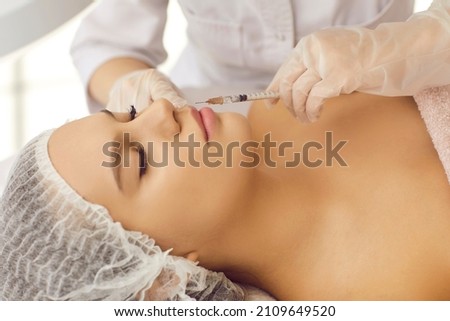 Beautiful young lady getting beauty treatment at clinic of aesthetic medicine and receiving injection for bigger, plumper lips. Doctor's hands in gloves holding syringe at woman's face, close up Royalty-Free Stock Photo #2109649520