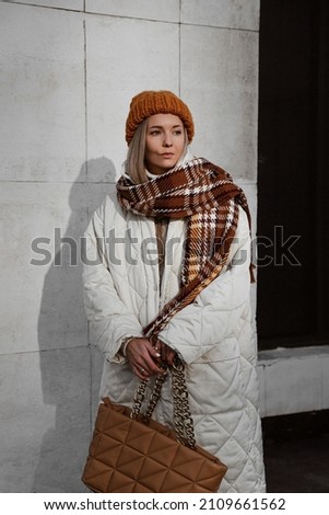 portrait of beautiful blonde girl dressed in white quilted down jacket, milky sweater and massive shoes, brown pants, knitted caramel hat scarf and bag, stylish trendy fashion outlook, lifestyle model