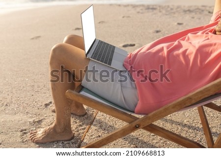 Low section of relaxed senior biracial man sitting with laptop on folding chair at beach. copy space, lifestyle and telecommunications. Royalty-Free Stock Photo #2109668813