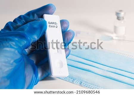 The doctor's hands with blue gloves hold an antigen test. On the table a blue surgical mask, coronavirus vaccine and vaccine vial syringe.