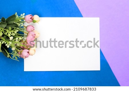 Blank paper card with artificial flowers on colorful background. Copy space for your text lay flat, top view