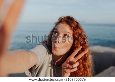 Carefree young woman taking a selfie while standing next to the sea. Young woman making a funny face and showing the peace sign. Cheerful young woman having fun in the summer sun.