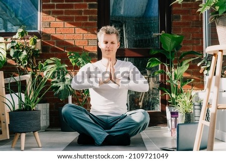 Young man doing yoga position Namaste in cozy loft style room full of home plants on sunny morning. Online class yoga practice. Healthy Lifestyle. Home online workout during a lockdown