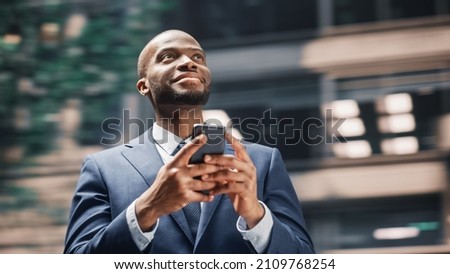 Street Shot: Portrait of Happy Black Businessman Using Smartphone in City. Smiling African American Entrepreneur Using Mobile Phone App Stock Market Investment. Blur Motion Background.