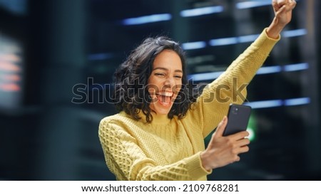 Street Shot: Portrait of Beautiful Latin Woman Using Smartphone, Celebrating Successful Victory with Yes Gesture. Smiling Hispanic Female Entrepreneur Using Mobile Phone Happily. Blur Motion Shot. Royalty-Free Stock Photo #2109768281
