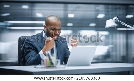 Office: Happy Successful Black Businessman Sitting at Desk Using Laptop Computer, Celebrate Success. Entrepreneur in Suit working with Stock Market App Smiles, Happy Victory. Motion Blur Background. Royalty-Free Stock Photo #2109768380