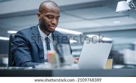 Office: Happy Successful Black Businessman Sitting at Desk Using Laptop Computer. African American Entrepreneur in Suit working with Stock Market Investing. Motion Blur Background.