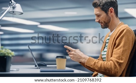 Modern Office: Businessman Sitting at His Desk Using Smartphone, Laptop Computer. Digital Entrepreneur working with Big Digital Data e-Commerce. Motion Blur Background Showing Active Work Day.