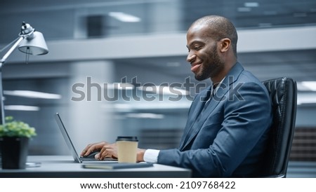 Modern Office: Successful Businessman Sitting at Desk Using Laptop Computer. African American Entrepreneur in Suit working with Stock Market Investing. Motion Blur Background Showing Active Work Day.