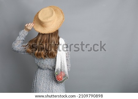 Attractive young girl in staw hat holding a mesh bag with vegetables. Picture taken from the back.Studio shot on grey background with copy space.

