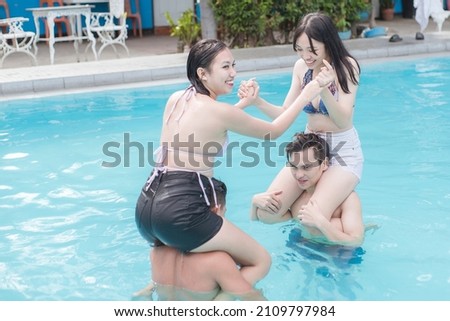 Teenage women in bikini tops and shorts sitting on their boyfriends shoulders and play fighting in a swimming pool. Fun chicken fights also known as shoulder wars.
