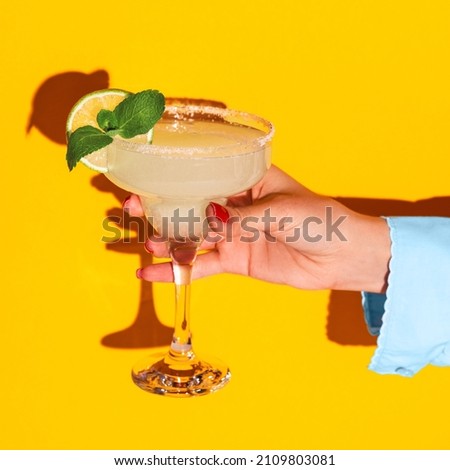 Sweet and sour. Female hand holding glass with margarita cocktail isolated on bright yellow neon background with shadow. Complementary colors, white, blue and yellow. Copy space for ad. Pop art Royalty-Free Stock Photo #2109803081
