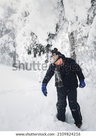 Boy in funny hat having fun in snow covered pine forest on Zlatibor mountain. Happy child wearing warm winter clothes getting snow shower under pine branch. Active kid spending wintertime outside.