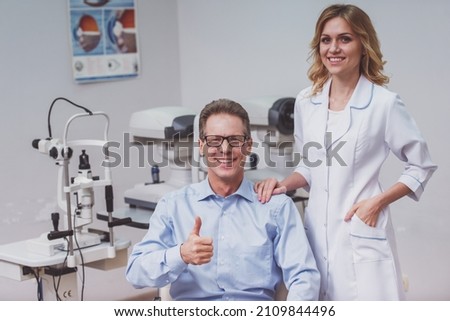 Handsome middle aged man at the ophthalmologist on consultation. Young beautiful female doctor standing near. Both smiling
