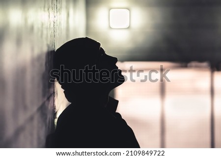 Serious sad man in dark. Depressed anxious person suffer from trauma, solitude or drug addiction. Homeless outcast with shame after mistake. Dramatic moody silhouette with melancholy, grief or sorrow. Royalty-Free Stock Photo #2109849722