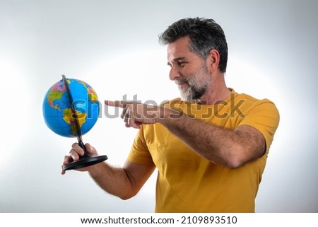 Mature bearded guy in casual t-shirt posing isolated on grey wall background studio portrait. People lifestyle concept. Mock up copy space. Pointing index finger on world globe