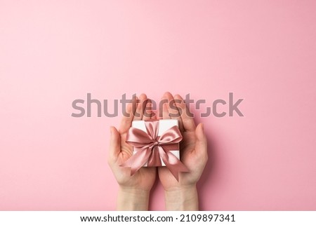 First person top view photo of valentine's day decorations female hands holding small white giftbox with pink ribbon bow on palms on isolated pastel pink background with empty space