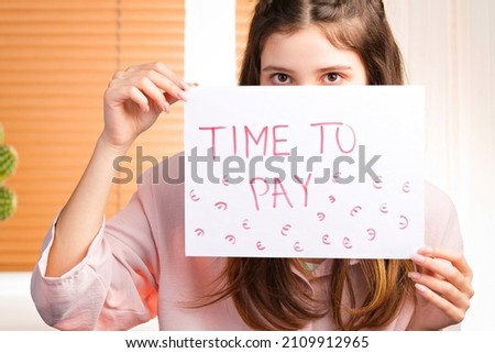 Close up photo of a young woman covering her face with a paper holding in her hands with an inscription of time to pay.