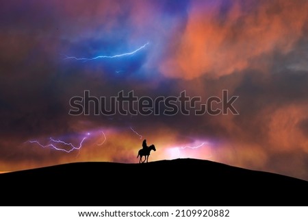 Silhouette of a lone rider on a horse standing on hill. Behind him is Dramatic storm sky and lighting behinde him the on the sky. Royalty-Free Stock Photo #2109920882