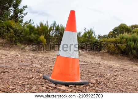 Photograph of an old and damaged orange safety cone with torn fluorescent tape on a rocky dirt road in front of trees and a cable fence