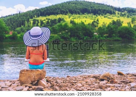 Rear view of young girl dressed in a straw beach sun hat working or studying remotely with a laptop outdoors on the river in front of the mountains. Copy space.