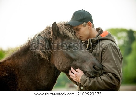 Young blonde caucasian man with cap petting a domesticated dark brown Icelandic mare. Human and animal equine friendship.  Royalty-Free Stock Photo #2109929573