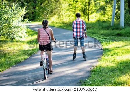Two friends riding a bicycle and roller skating in a park in summer. 