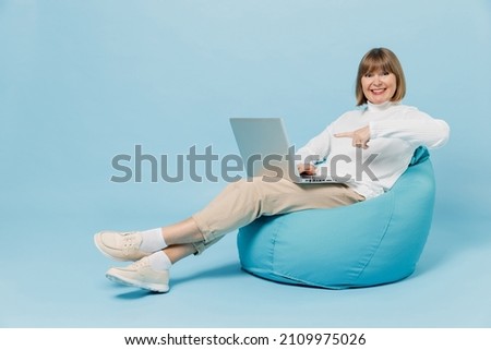 Full body elderly smiling woman 50s in white knitted sweater sit in bag chair hold use work point on laptop pc computer isolated on plain blue color background studio portrait People lifestyle concept