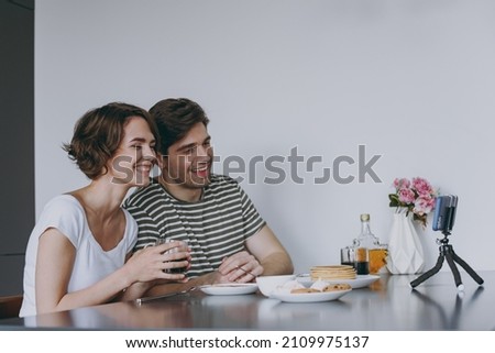 Young happy smiling couple two woman man 20s in casual clothes sit by table eat pancake talk video call mobile cell phone drink coffee cook food in light kitchen at home together Healthy diet concept