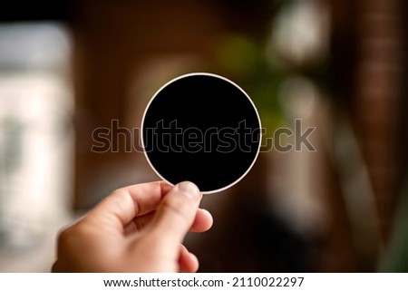 Hand holding a circle-shaped black bumper sticker that is blank for text space and copy space with plenty negative space to place a logo or text. Brick background with plants, windows, natural light. Royalty-Free Stock Photo #2110022297