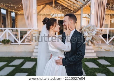 A young groom and a beautiful, smiling brunette bride in a white dress stand embracing on the green grass, in nature, against the backdrop of a cottage, at home. Wedding portrait, photography.